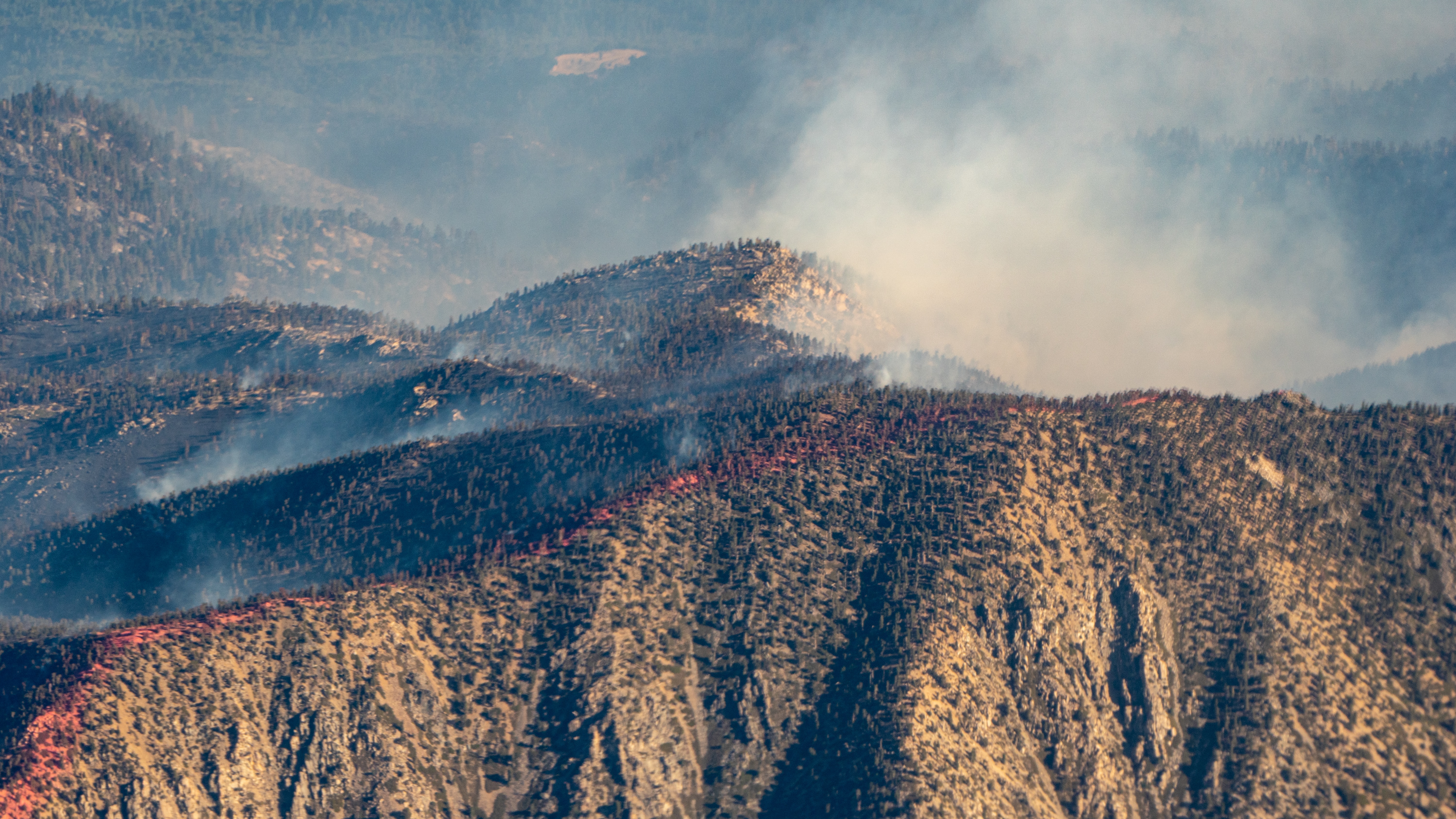 Wildfire smoke rises above dry and scorched mountaintops. Red fire retardant lines the ridgeline of the mountain in the foreground.