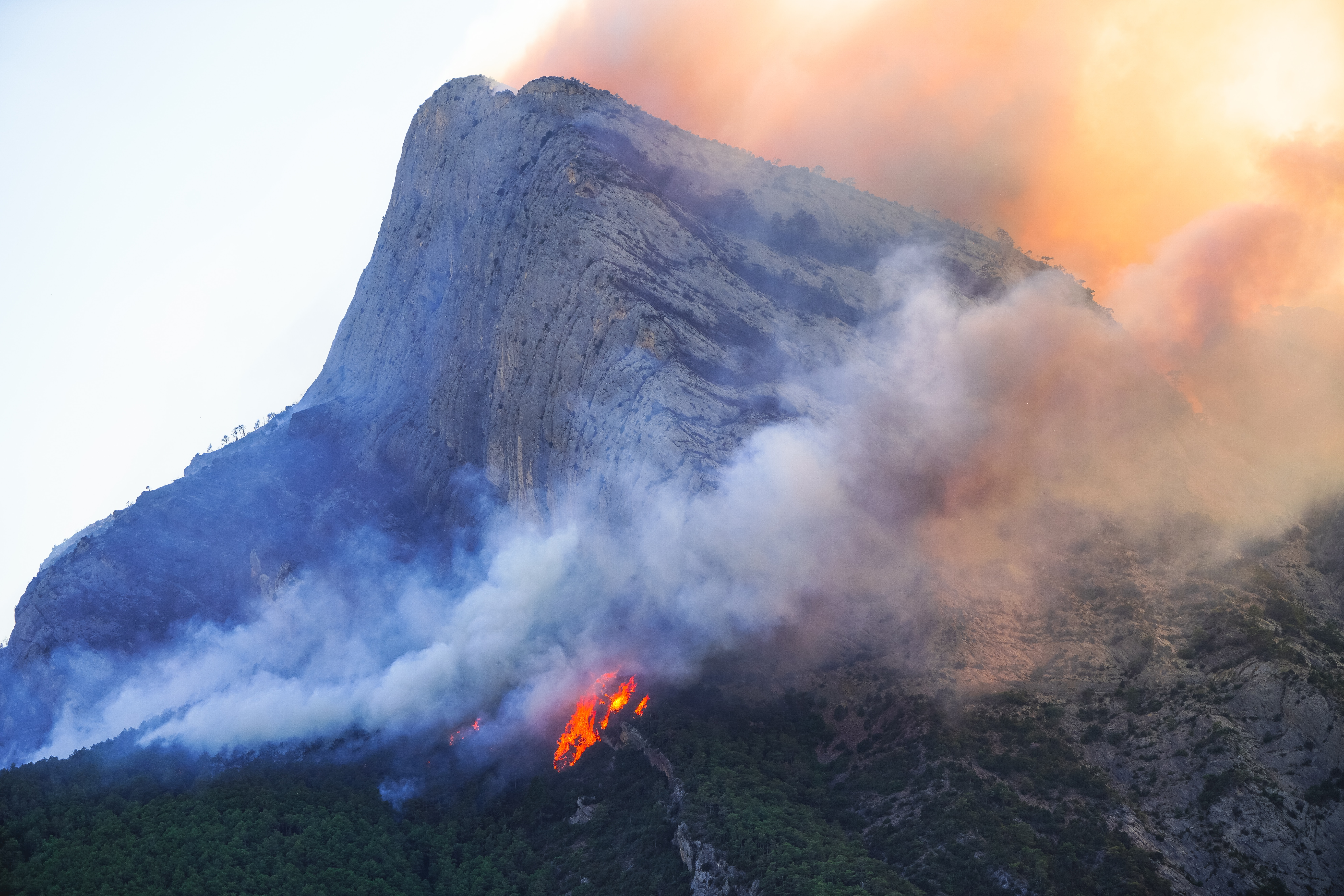 A wildfire burns vegetation beneath a rocky mountain peak.